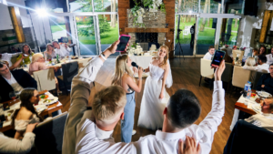 Chrzestny w towarzystwie innych gości weselnych, śmiejących się i rozmawiających, celebruje radosne chwile na weselu.